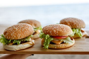 Four classic beef burgers with fresh lettuce and ripe tomatoes, served on a wooden cutting board against a blurred beach background