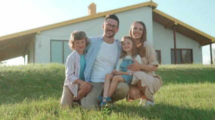 Caucasian family sitting together in front of their large house in sunny day. Parents together with son and daughter smiling as group and looking directly at camera. Lovely family moment.