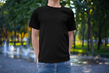 Mockup of a casual black cotton t-shirt on a guy with hands in pockets, on the background of a park, fountain, trees, front view.