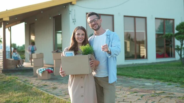 Caucasian couple standing in front of newly purchased house. Woman holding boxes of their belongings. Man waving keys to house to camera. Children playing in background. Moving to new home.