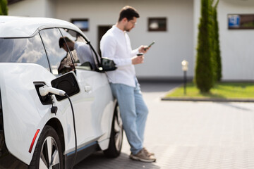 Man Charging Electric Car While Using Smartphone Outdoors