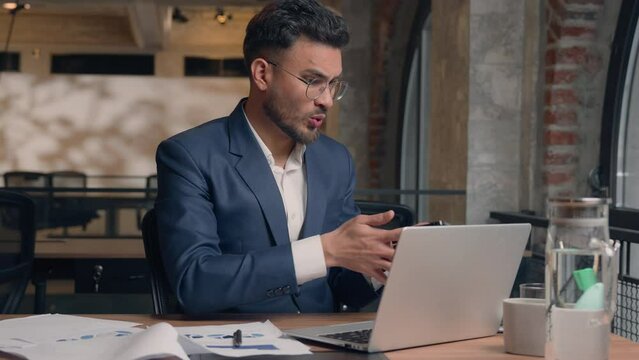 Arabian business male guy businessman in office busy stressed annoyed anxious Indian man answer call talk mobile phone consult search data documents typing laptop difficult multitasking work overwork