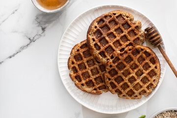 Homemade oatmeal and chia seed waffles on a white plate, accompanied by honey and ingredients