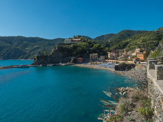 Fototapeta premium Picturesque view of Monterosso al Mare with beach, colorful houses, train rails, bell tower and Torre Bastioni with green cliffs, clear blue sky and sea. National park Cinque Terre, Liguria, Italy