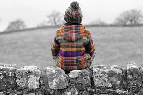 Child in Plaid Jacket Sitting on Stone Wall in Rural Landscape