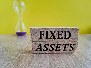 Concept words fixed assets on beautiful brick blocks. Beautiful yellow background, hourglass. Business and fixed assets concept.