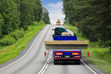 Heavy truck and trailer transporting wide load along highway with pilot vehicle ahead leading the way. Rear view, copy space.