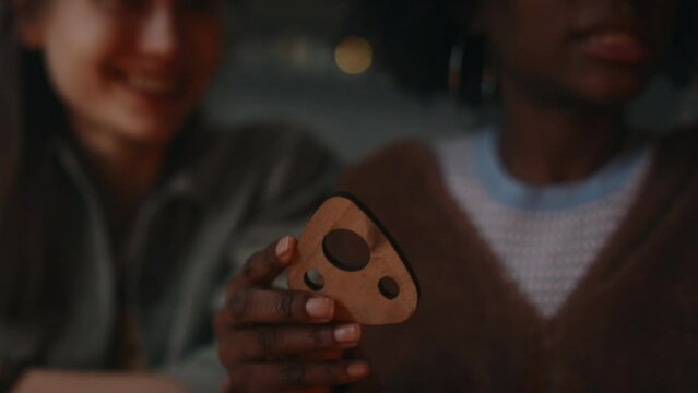Cropped shot of unidentified girl holding planchette and explaining to friends how to use it during spiritualistic seance with talking board at night indoors, focus on planchette, blurred background