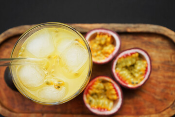 Passion fruits cut in half next to a glass of juice with ice on a wooden tray on a dark background