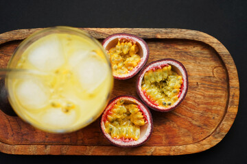 Passion fruits cut in half next to a glass of juice with ice on a wooden tray on a dark background