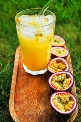 Passion fruits cut in half next to a glass of juice with ice on a wooden tray against a background of green grass