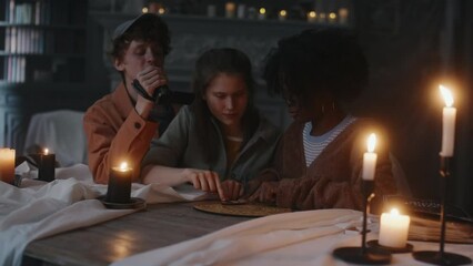 Medium shot of diverse adolescents having fun while using talking board at table by candlelight and guy recording video on camera. Evoking evil spirit who blowing out candles and frightening kids