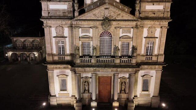 A dramatic night aerial shot starting from the base of the Sanctuary of Bom Jesus do Monte in Braga, Portugal, ascending to reveal the brilliantly lit rooftop and architectural details glowing against