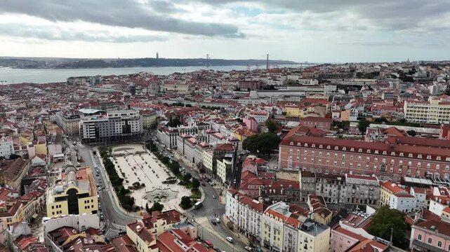 Drone flies over Lisbon toward the Tagus and 25 de Abril Bridge, passing Rossio Square and iconic landmarks in a sweeping panoramic cityscape view