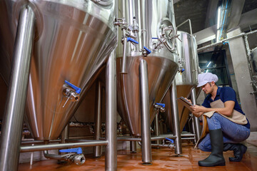 A young Asian male brewer holds a tablet in his hand and is inspecting craft beer to check the flavor and color of the beer in the brewery industry.