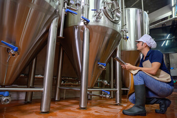 A young male brewer holds a tablet in his hand and is inspecting craft beer to check the flavor and color of the beer in the brewery industry.