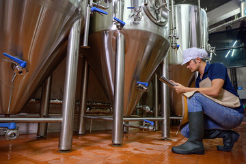 A young male brewer holds a tablet in his hand and is inspecting craft beer to check the flavor and color of the beer in the brewery industry.