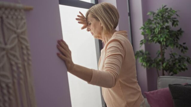An elderly blonde woman leaning against a window in her living room, appearing dizzy in a modern apartment with purple walls and indoor plants.