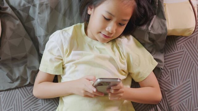 Top view of adorable Asian kid girl, lied in bed, enjoys her smartphone in a sunlit room. Her expression hints at digital fun, highlighting the allure of home tech, playing game and e-learning.