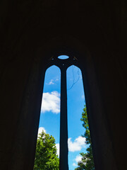 View of the blue sky with green trees through the window of the old temple. A broken window of a small temple