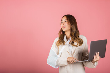 Asian business woman confident smiling broken arm after accident and wear arm splint for treatment but still have to work she hold laptop computer studio isolated on pink background, copy space