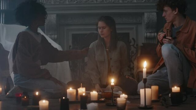 Full shot of frightened teenagers watching their friend being possessed by devil while performing ceremony by candlelight in abandoned house at night