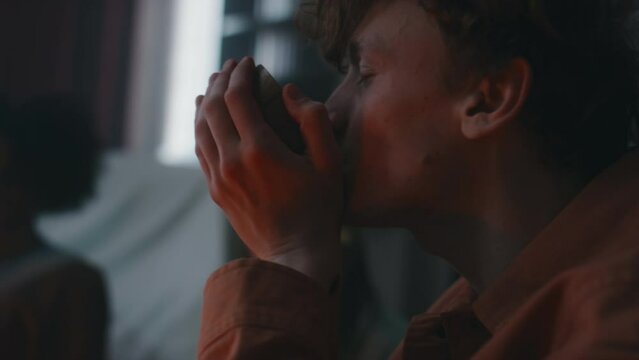 Side closeup of Caucasian teenage guy drinking blood from wooden ritual cup during ceremony for summoning satanic beings with friends in house at night
