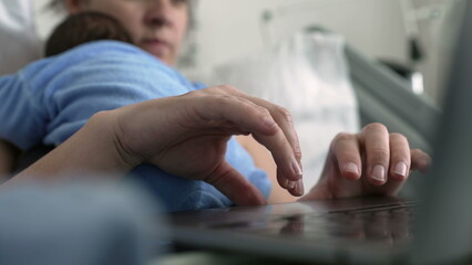 Hands of new mother browsing internet on laptop while holding sleeping newborn baby, multitasking in hospital bed, combining parenting and work, postnatal care