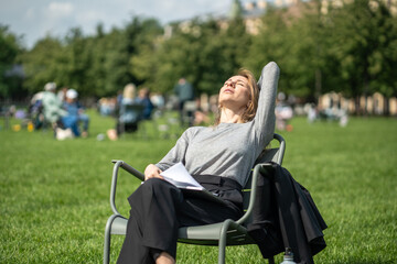 Relaxed woman sits on chair outdoor enjoying fresh air in public place with closed eyes. Calm...