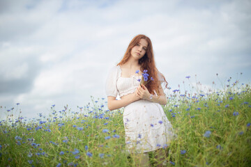 A girl with long curly red hair in white clothes stands in a meadow with cornflowers, holding a bouquet of cornflowers in her hands, summer day, cloudy warm weather
