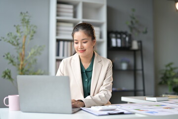 Young asian businesswoman multitasks on her laptop in a modern office, exuding confidence and professionalism surrounded by paperwork, charts, and graphs