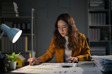 Determined businesswoman works late in an office, using a calculator and jotting down notes on...