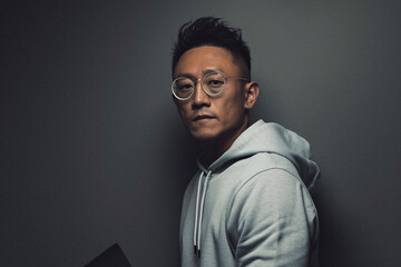 A person wearing glasses and a hoodie stands against a neutral background, looking intently forward, their expression serious and thoughtful, captured in soft, moody lighting.