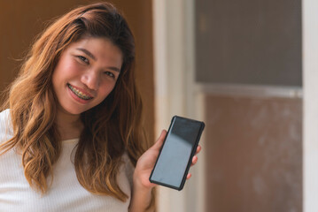 Asian woman in her 30s standing outside her apartment flat, showing off a cellphone app. Blank phone screen for editing.