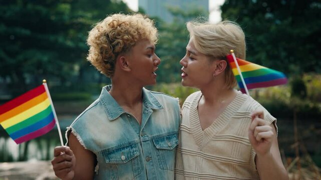 Positive young gays with rainbow flags kiss and smiles standing against park pond. Smiling Asian men from local LGBT community celebrate pride month. Family of Korean guys shows love