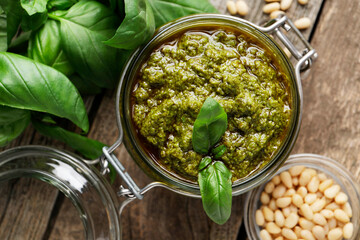 Tasty pesto sauce in jar, pine nuts and basil on wooden table, top view