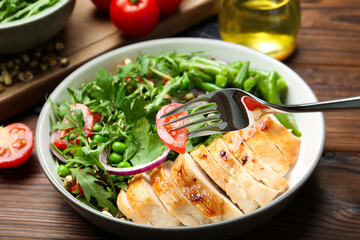 Healthy meal. Eating tasty salad and chicken breast with fork from bowl on wooden table, closeup
