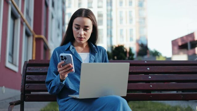 A young woman wearing a blue shirt dress sits on a bench while working remotely on her laptop and phone.