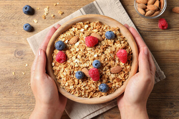Woman holding bowl of tasty granola with berries and nuts at wooden table, top view