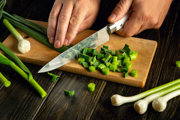 A cook slices young green garlic stalks with a knife on a wooden cutting board to prepare dinner.