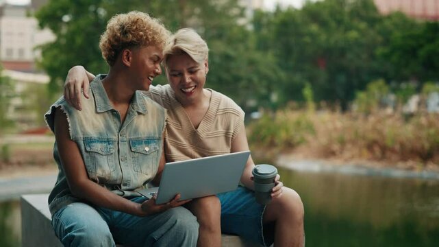 Cheerful young gay men with laptop rest on pond bank in urban park slow motion. Homosexual male partners watch video online via computer outdoors. Pride month in local LGBTQI community