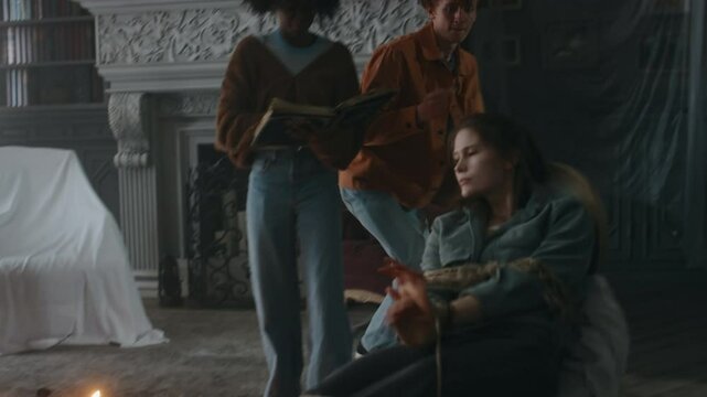 Medium long shot of lady reading prayers in Latin from occult book and guy lighting palo santo stick to exorcise devils from friends body, drawing cross with embers on writhing girls forehead indoors