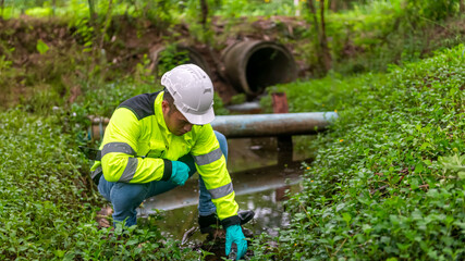 An ecologist in a green jacket sitting aside water try to scoop the water in a glass tube to...