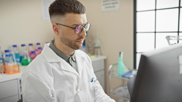 A focused hispanic man in a lab coat analyzes data on a computer screen at a bright laboratory setting.