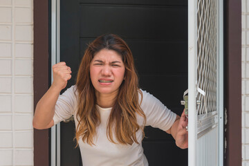 An angry asian woman in her 30s stands just outside her apartment flat, looking livid and holding...