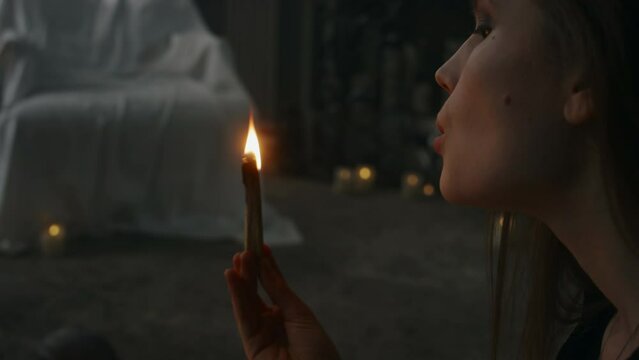 Side tilt shot of young witch sitting among burning candles and lighting palo santo wood stick, blowing it out to clear house from negative energy and prepare room for dark rituals