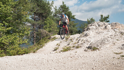 Obraz premium Mountain bike rider riding an electric bike along a white gravel trail on a sunny summer day. Outdoor sports activity concept.