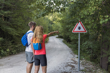 Man and woman pointing fingers in the distance while hiking near the woods with a tick warning sign. Risk of tick-borne disease concept.