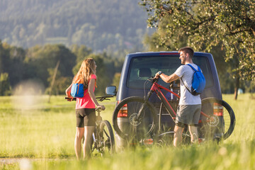Young man and a woman preparing for off-road biking, taking down electric mountain bikes from the bike rack on the camper van. © 24K-Production