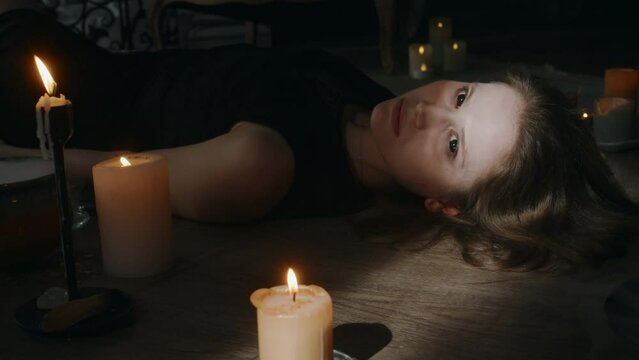 Side shot of young Caucasian lady in black dress possessed by devil waking up lying on wooden floor midst burning candles